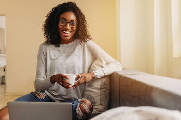 Lächelnde Frau in modischen zerrissenen Jeans hält eine Kaffeetasse, während sie zu Hause am Laptop arbeitet. Geschäftsfrau sitzt zu Hause auf dem Sofa und arbeitet am Laptop. - JLPSF01720