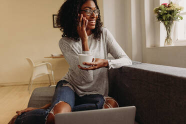 Businesswoman sitting on sofa at home and talking over mobile phone while working on laptop. Smiling woman in fashionable torn jeans holding a coffee cup while working on laptop computer at home. - JLPSF01721