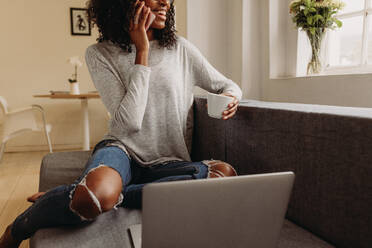 Businesswoman sitting on sofa at home and talking over mobile phone while working on laptop. Smiling woman in fashionable torn jeans holding a coffee cup while working on laptop computer at home. - JLPSF01722