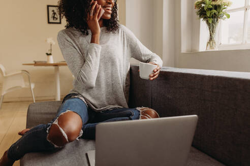 Geschäftsfrau sitzt auf dem Sofa zu Hause und spricht über Handy, während sie an einem Laptop arbeitet. Lächelnde Frau in modischen zerrissenen Jeans hält eine Kaffeetasse, während sie zu Hause an einem Laptop arbeitet. - JLPSF01722