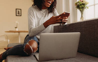 Businesswoman sitting on sofa at home using mobile phone while working on laptop. Smiling woman in fashionable torn jeans holding a mobile phone while working on laptop computer at home. - JLPSF01723