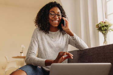 Businesswoman sitting on sofa at home and talking over mobile phone while working on laptop. Smiling woman in fashionable torn jeans working on laptop computer at home and talking on mobile phone. - JLPSF01724