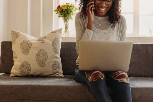 Geschäftsfrau sitzt auf dem Sofa zu Hause und spricht über das Mobiltelefon, während sie an einem Laptop arbeitet. Lächelnde Frau in modischen zerrissenen Jeans spricht über das Mobiltelefon, während sie zu Hause an einem Laptop arbeitet. - JLPSF01727