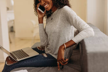 Businesswoman sitting on sofa at home talking over mobile phone while working on laptop. Woman entrepreneur in fashionable torn jeans speaking on mobile phone with laptop computer on her lap. - JLPSF01730