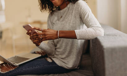 Businesswoman sitting on sofa at home using mobile phone while working on laptop. Woman working on laptop computer at home and managing her business. - JLPSF01731