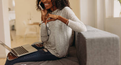 Woman entrepreneur in fashionable torn jeans speaking on mobile phone with laptop computer on her lap. Businesswoman sitting on sofa at home talking over mobile phone holding her spectacles in hand while working on laptop. - JLPSF01732