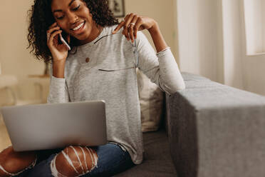 Woman entrepreneur in fashionable torn jeans speaking on mobile phone with laptop computer on her lap. Businesswoman sitting on sofa at home talking over mobile phone holding her spectacles in hand while working on laptop. - JLPSF01733