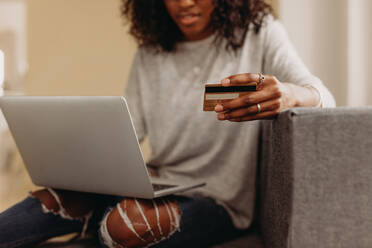 Frau in modischen zerrissenen Jeans sitzt auf dem Sofa zu Hause hält eine Kreditkarte mit einem Laptop auf dem Schoß. Frau macht Online-Zahlung mit Kreditkarte auf ihrem Laptop-Computer. - JLPSF01734