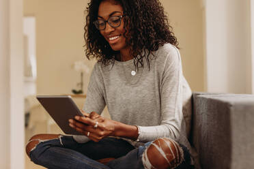 Frau sitzt zu Hause auf dem Sofa und arbeitet an einem Tablet-PC. Lächelnde Frau, die einen Tablet-PC benutzt, während sie zu Hause mit gekreuzten Beinen sitzt. - JLPSF01737