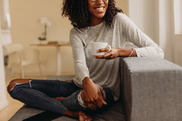 Smiling woman in fashionable torn jeans holding a coffee cup relaxing at home. Woman sitting with legs crossed on sofa at home drinking coffee and looking away. - JLPSF01738