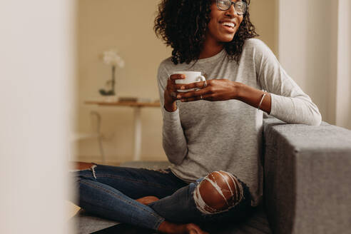 Lächelnde Frau in modischen zerrissenen Jeans, die eine Kaffeetasse hält und sich zu Hause entspannt. Frau sitzt mit gekreuzten Beinen auf dem Sofa zu Hause, trinkt Kaffee und schaut weg. - JLPSF01739