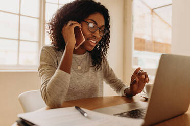 Woman entrepreneur managing her business from home working on laptop computer and discussing business over mobile phone. Businesswoman sitting at home talking on mobile phone while working on laptop. - JLPSF01746