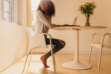 Lächelnde Geschäftsfrau in modischen Jeans, die zu Hause an einem runden Tisch sitzt und an einem Laptop arbeitet und zurückblickt. - JLPSF01748