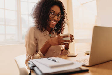 Businesswoman working on laptop computer sitting at home holding a coffee cup in hand. Smiling woman entrepreneur sitting at home enjoying coffee while working on laptop. - JLPSF01758