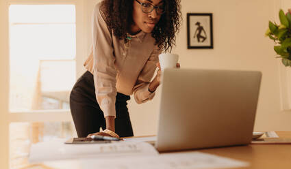 Businesswoman looking at laptop computer holding a coffee cup in hand. Woman entrepreneur working on laptop from home standing beside the table. - JLPSF01766