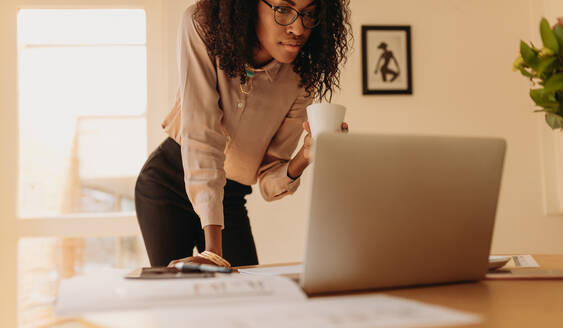 Geschäftsfrau, die auf einen Laptop schaut und eine Kaffeetasse in der Hand hält. Unternehmerin, die von zu Hause aus am Laptop arbeitet und neben dem Tisch steht. - JLPSF01766