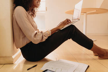 Side view of a woman entrepreneur in formal attire working on laptop computer sitting at home. Businesswoman sitting on floor at home working on laptop with business papers by her side. - JLPSF01771