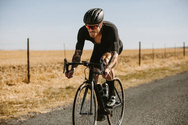 Entschlossener Radfahrer, der auf einer Landstraße Fahrrad fährt. Profi-Radfahrer mit Helm und Sonnenbrille beim Training im Freien. - JLPSF01826