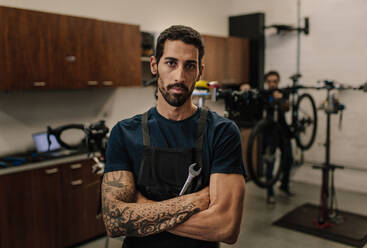 Worker standing with folded arms holding a spanner in workshop. Mechanics assembling bicycles in repair shop. - JLPSF01849