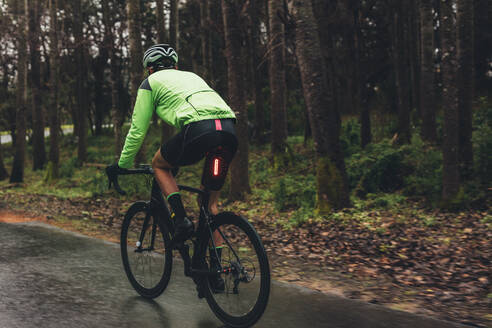 Männlicher Radfahrer auf einer Landstraße, der für einen Wettkampf trainiert. Ein Mann fährt mit dem Fahrrad auf einer nassen Straße durch den Wald. - JLPSF01889
