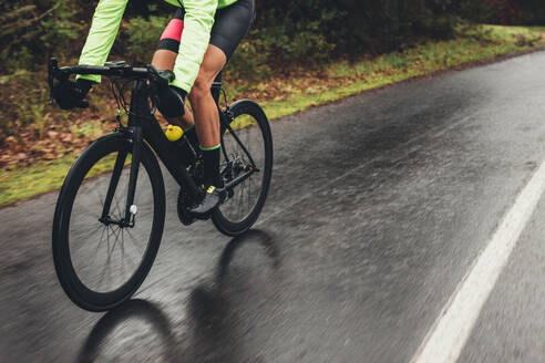 Männlicher Sportler in Radfahrerkleidung fährt auf nasser Straße Rad. Tiefschnittaufnahme eines Radfahrers beim Training im Freien an einem regnerischen Tag. - JLPSF01893