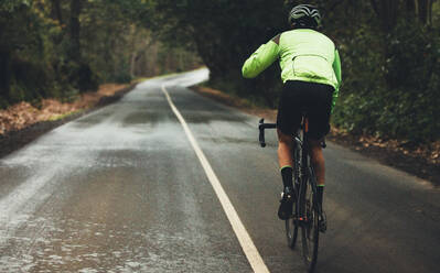 Rückansicht eines männlichen Sportlers, der an einem regnerischen Tag auf einer Landstraße radelt. Ein professioneller Radfahrer fährt auf einer leeren Landstraße durch den Wald. - JLPSF01899