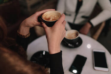 Nahaufnahme einer Frau, die eine Tasse Kaffee in den Händen hält, während sie mit einem Freund im Café sitzt. Weibliche Freunde trinken Kaffee im Café. - JLPSF01902