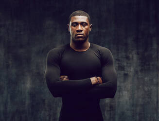 Muskulöser Mann in engem schwarzen Outfit steht mit verschränkten Armen im Fitnessstudio. - JLPSF01904