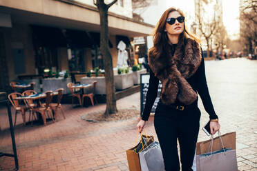 Fashionable young woman walking on the city street with shopping bags. Female model outdoors on road carrying shopping bags. - JLPSF01915