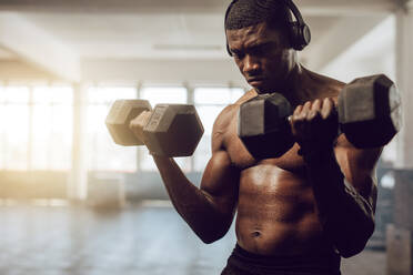 Ein Mann mit nacktem Oberkörper hört Musik beim Training im Fitnessstudio. Ein sportlicher Mann hebt Hanteln und trägt drahtlose Kopfhörer. - JLPSF01922