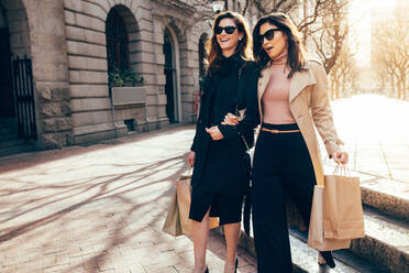 Stylish women friends walking on the city street with shopping bags. Female shoppers carrying shopping bags while walking along the road. - JLPSF01924