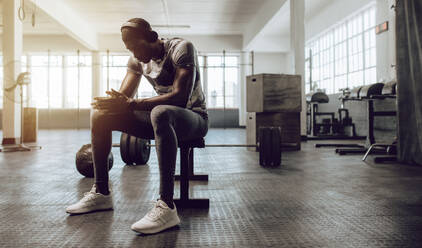 Crossfit guy relaxing after workout sitting on a bench in the gym. Man listening to music using wireless headphones and relaxing at the gym. - JLPSF01932