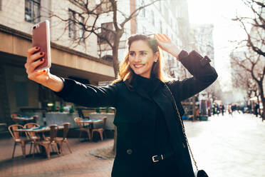 Beautiful young woman taking selfie using smart phone on the city street. Caucasian female model taking self portrait on the road. - JLPSF01937