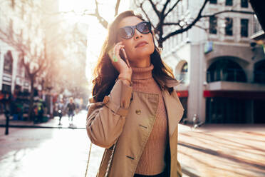 Schöne asiatische Frau zu Fuß entlang der Straße in der Stadt und sprechen auf dem Handy. Modische weibliche Modell mit Sonnenbrille zu Fuß auf der Straße der Stadt und mit Telefon. - JLPSF01938