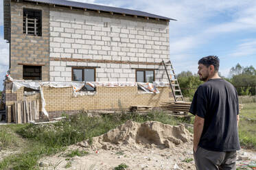 Mann steht an einem sonnigen Tag vor einem im Bau befindlichen Haus - ANAF00062