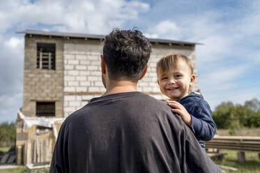 Father carrying son in front of house at construction site on sunny day - ANAF00063