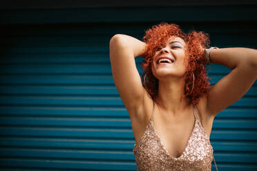 Woman in happy mood laughing with eyes closed holding her curly golden brown hair. Close up of a young woman in a glamorous dress outdoors. - JLPSF01943