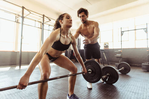 Personal Trainer unterstützt eine junge Frau beim Heben einer schweren Langhantel beim Training in einem Fitnessstudio. Fitte junge Frau hebt eine Langhantel mit Unterstützung eines männlichen Trainers - JLPSF01947