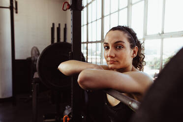 Junge Frau lehnt sich nach dem Training im Fitnessstudio über eine Langhantel. Frau ruht sich nach dem Training im Fitnessstudio aus. - JLPSF01948