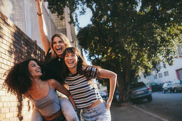 Außenaufnahme von multiethnischen Freundinnen, die draußen in der Stadt spazieren gehen und Spaß haben. Drei junge Frauen, die zusammen spazieren gehen, Huckepack nehmen und Spaß haben. - JLPSF01959