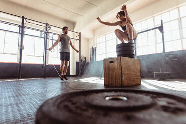 Ein Mann trainiert mit einem Springseil und eine Frau springt auf einer Box im Fitnessstudio. Ein sportliches Paar trainiert hart im Cross Fit Fitnessstudio. - JLPSF02021