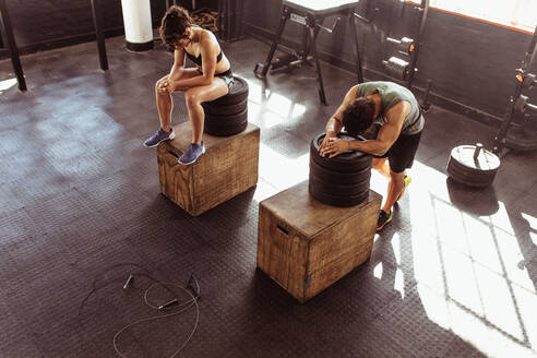 Fitness-Mann und Frau, die sich nach Übungen im Cross-Training-Fitnessstudio auf einer Box ausruhen. Junge Leute im Fitnessstudio, die sich nach einem anstrengenden Training ausruhen. Sie machen eine Pause nach einer intensiven körperlichen Trainingseinheit. - JLPSF02023