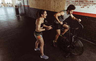 Personal Trainer motiviert Mann, der im Fitnessstudio auf dem Airbike trainiert. Mann macht intensives Training auf dem Fitnessrad mit weiblichem Trainer. - JLPSF02028