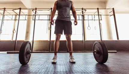 Mann mit Langhantel im Fitnessstudio. Männlicher Sportler vor dem Training mit schweren Gewichten im Fitnessstudio. Niedriger Winkel mit Fokus auf die Füße. - JLPSF02036