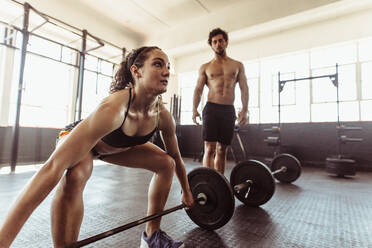 Eine fitte junge Frau trainiert mit einer Langhantel und ein Mann steht im Hintergrund. Eine Sportlerin hebt schwere Gewichte in einem Cross-Trainingsstudio. - JLPSF02051