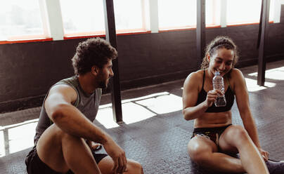 Junge Leute sitzen auf dem Boden und entspannen sich in der Turnhalle nach dem Training mit Frau trinken Wasser. Paar entspannt nach dem Training im Fitnessstudio. - JLPSF02059