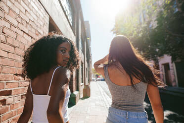 Rückansicht einer jungen afrikanischen Frau, die über ihre Schulter schaut, während sie mit ihrer Freundin auf einer Straße in der Stadt spazieren geht. - JLPSF02061