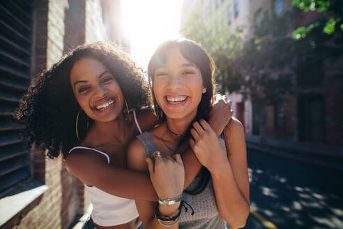 Porträt von zwei jungen Frauen auf einer Straße in der Stadt, die sich amüsieren. Freundinnen auf der Straße, die sich umarmen und lächeln. - JLPSF02066