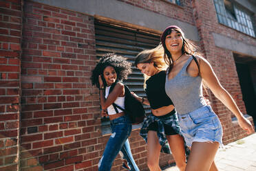 Eine gemischtrassige Gruppe von Freunden geht eine Straße entlang. Drei junge Frauen gehen im Freien auf der Straße. - JLPSF02068