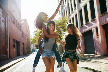 Außenaufnahme einer jungen Frau, die ihre Freundin auf dem Rücken trägt. Drei junge Frauen amüsieren sich auf der Straße. - JLPSF02074